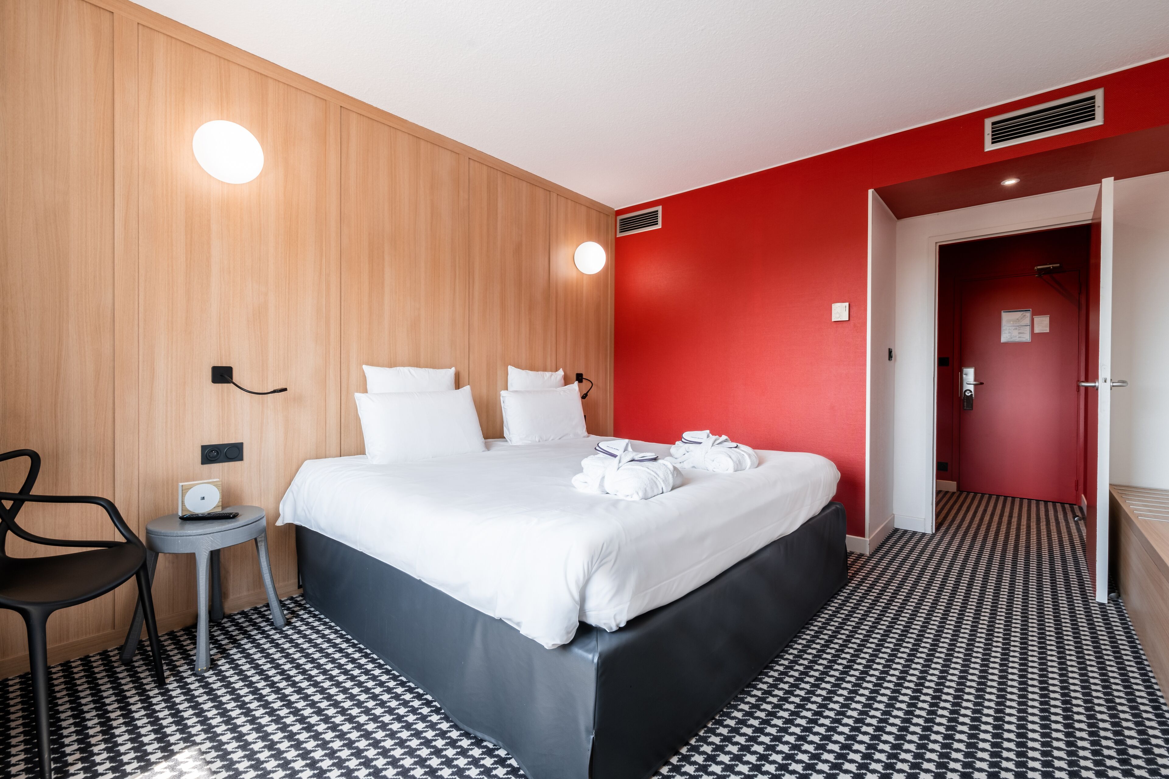 A hotel room with a wooden panel wall, a red accent wall, a black and white patterned floor, a bed with white linens, a small round table with a lamp, and a chair.
