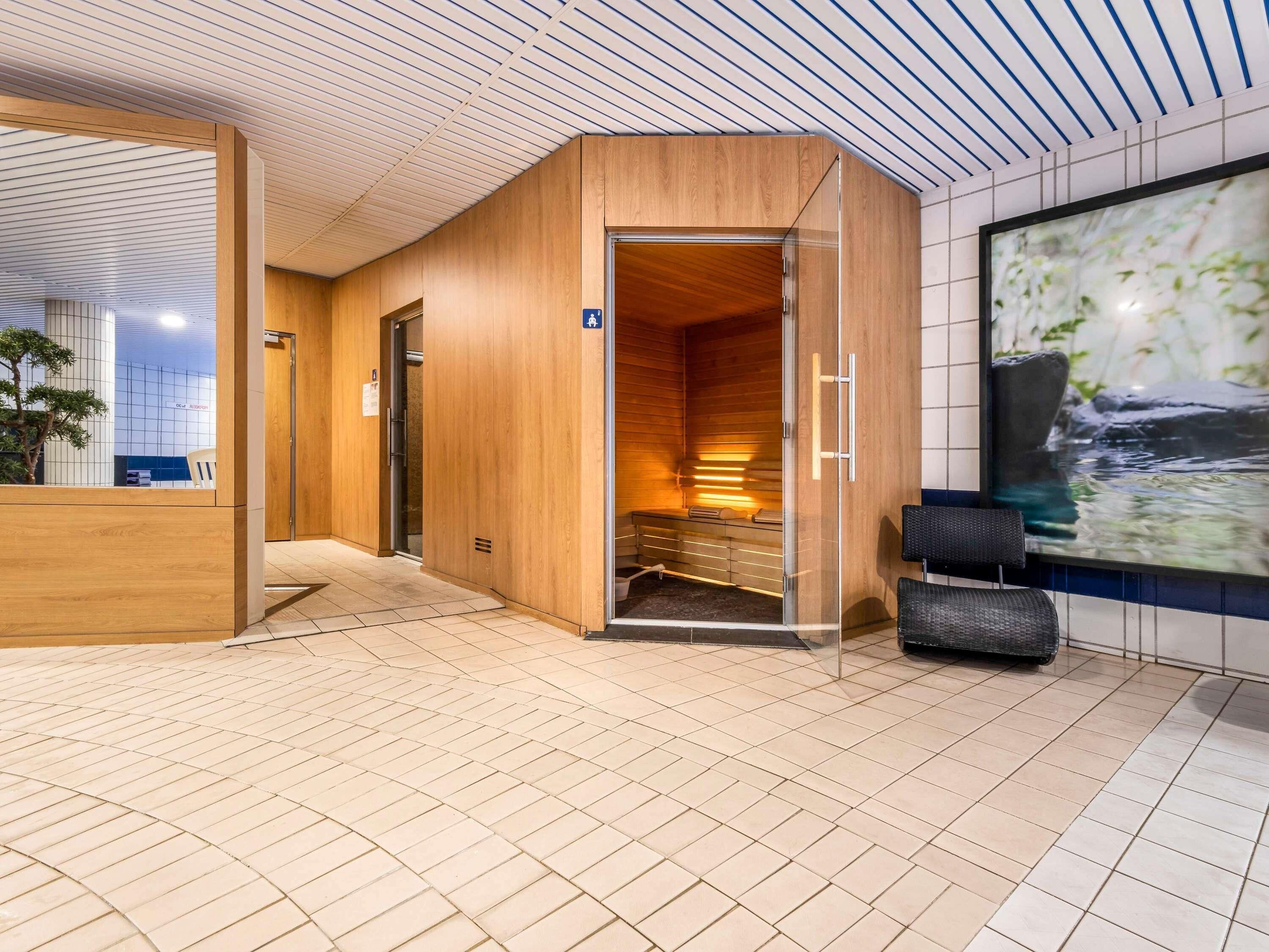 Una sauna moderna en el interior con paredes de madera y puerta de cristal, un área de recepción con una planta y un ventanal con vistas a la naturaleza.