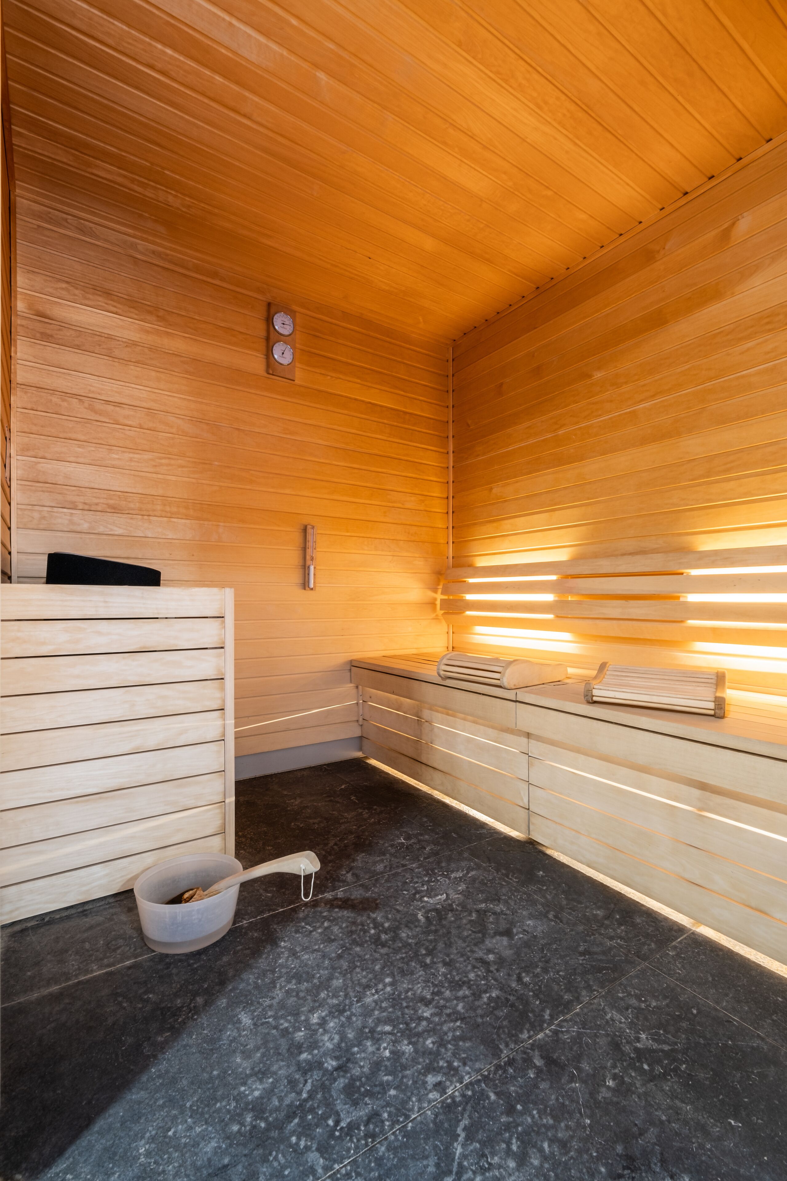 Una sala de sauna con paredes de madera y un banco. Hay un perchero y un cubo con un palo de madera.