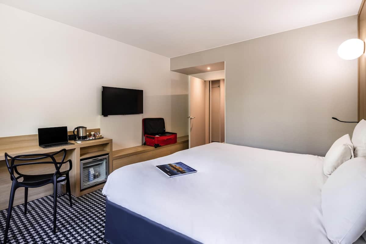 A hotel room with a bed, a desk with a laptop, a television, a red suitcase, and a small refrigerator.
