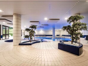 A modern indoor swimming pool area with tiled flooring, seating by the pool, and potted trees. - Mercure Tours Sud (Joue-les-Tours)