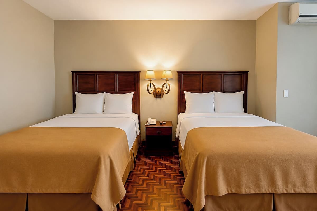 A hotel room with two beds, a nightstand with a lamp, and a wooden herringbone floor.