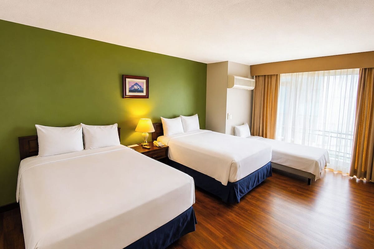 A hotel room with two beds, a bedside table with a lamp, a picture on the wall, and a large window with curtains.
