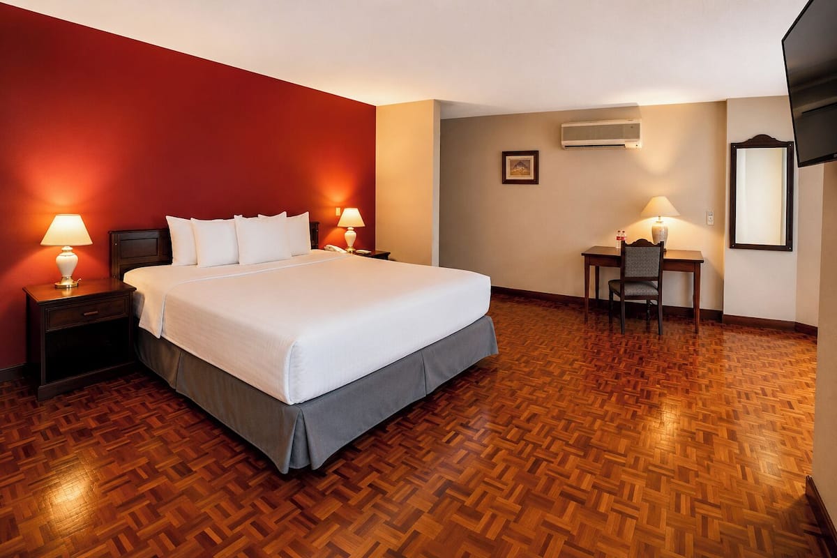A hotel room with a bed, nightstands, a desk with a chair, an air conditioning unit, and a wall-mounted television.
