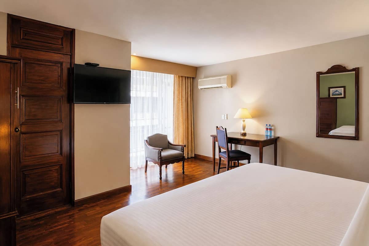 A hotel room with a bed, a desk with a chair, a TV, an air conditioner, and a mirror.