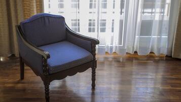 A chair with a woven backrest and armrests, positioned in a room with wooden flooring and a large window with sheer curtains.