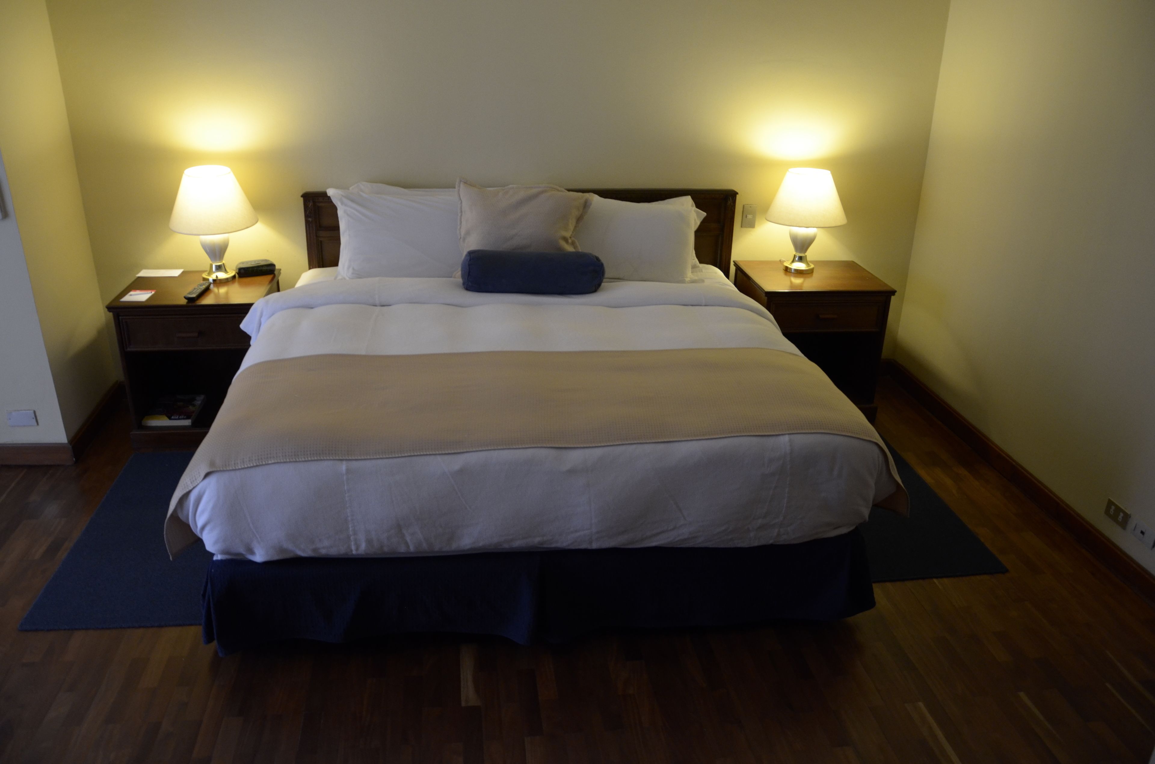 A neatly made bed with white linens, two bedside tables with lamps, and a dark-colored rug on a wooden floor.