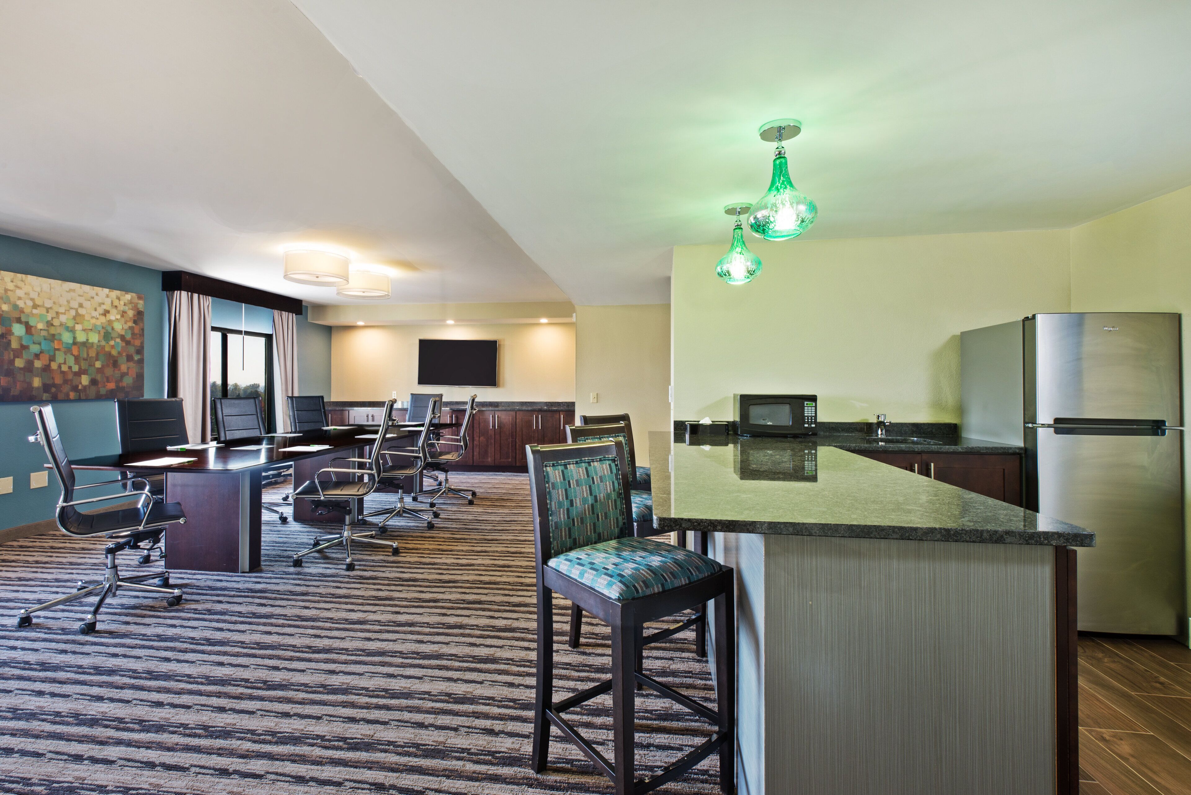 A modern hotel room with a kitchen area, a dining table with chairs, and a large window.