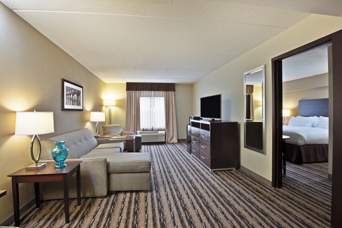 A hotel room with a sofa, a desk with a lamp, a television, a dresser, and a bed.