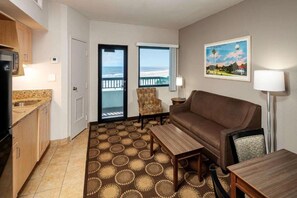 Habitación de hotel con kitchenette, sofá, mesa de comedor, alfombra estampada y vista a la playa desde la puerta.