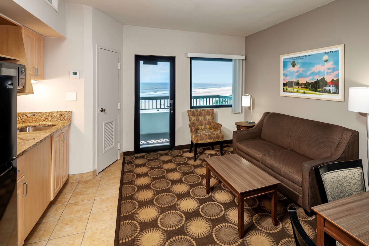Une chambre d’hôtel compacte comprenant un coin cuisine, un canapé, une table à manger et une chaise. Une grande fenêtre offre une vue sur la plage, et un tableau encadré orne le mur.