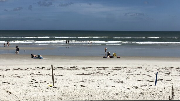 Une plage où l’on voit des gens se promener et s’asseoir sur le sable, un ciel dégagé et l’océan en arrière-plan.