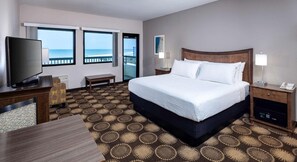 A hotel room with a large bed, a TV, a desk, and a view of the ocean.