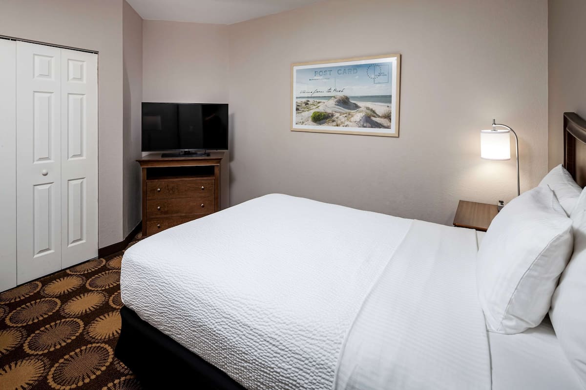 Une chambre d’hôtel avec un lit, un téléviseur posé sur un meuble en bois, un tableau encadré accroché au mur et une table de chevet avec une lampe.
