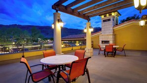 Une terrasse extérieure avec des tables rondes et des chaises orange, une cheminée en pierre et une vue sur le paysage urbain au crépuscule.
