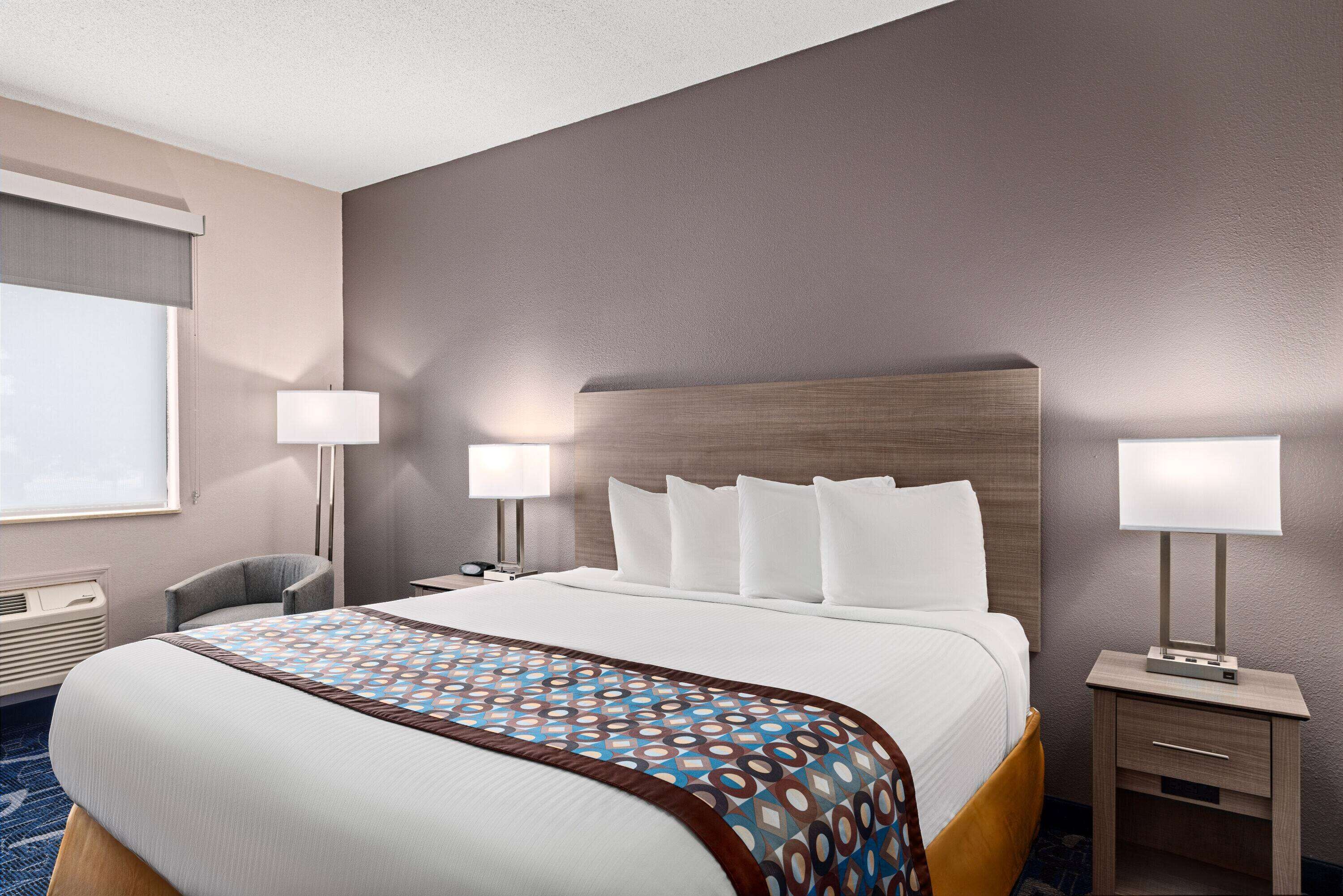 A hotel room with a large bed, two bedside tables with lamps, a chair, and a window with blinds.