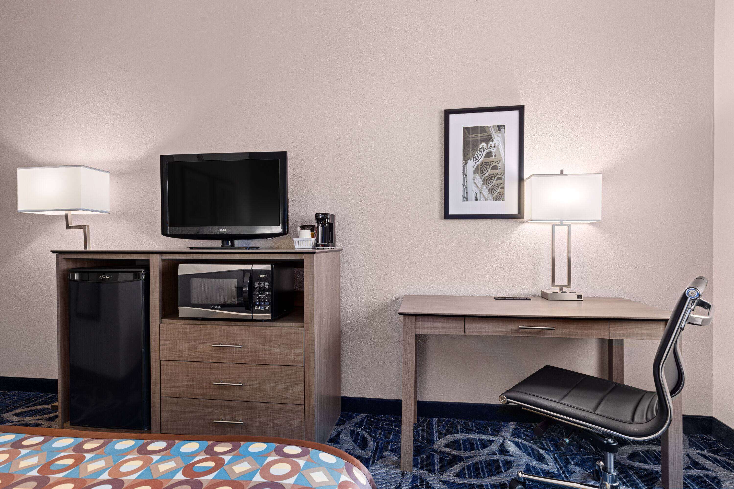 Habitación de hotel con cama, escritorio con silla, televisor, microondas, refrigerador y un cuadro en la pared.