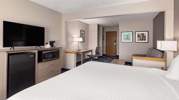 A hotel room with a large bed, a flat-screen TV, a refrigerator, a microwave, a desk with a chair, and a small table with a lamp.