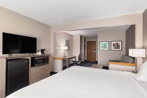 Ein Hotelzimmer mit einem großen Bett, einem Flachbildfernseher, einem Kühlschrank, einer Mikrowelle, einem Schreibtisch mit Stuhl und einem kleinen Tisch mit Lampe.