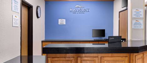Ein Empfangsbereich mit einem Schild von Baymont Inn & Suites, einer Uhr, einem Fernseher und einem Check-in-Terminal.