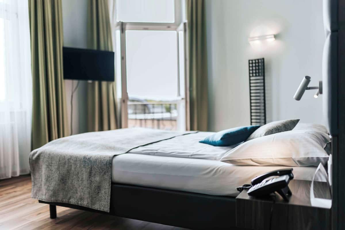 Uma cama bem arrumada com manta e travesseiros cinzas, um telefone de mesa e um aplique na parede.