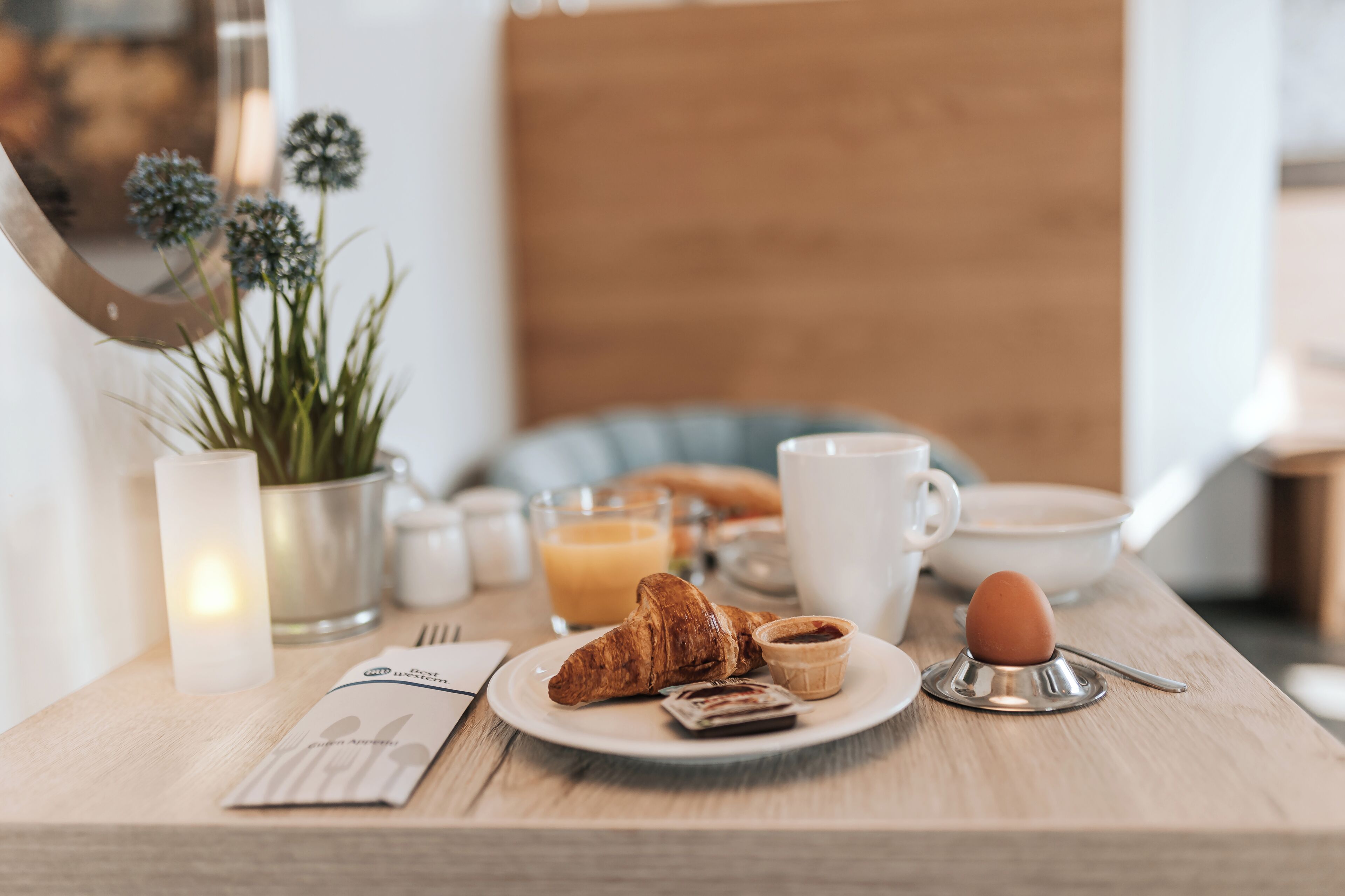 Desayuno con cruasanes, una taza de café, un plato con un cruasán y un frasco pequeño de mermelada, una vela, un vaso de jugo de naranja y una planta al fondo.