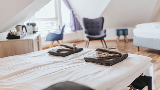A bedroom with a bed, two chairs, a small table with a kettle, and a window with curtains.