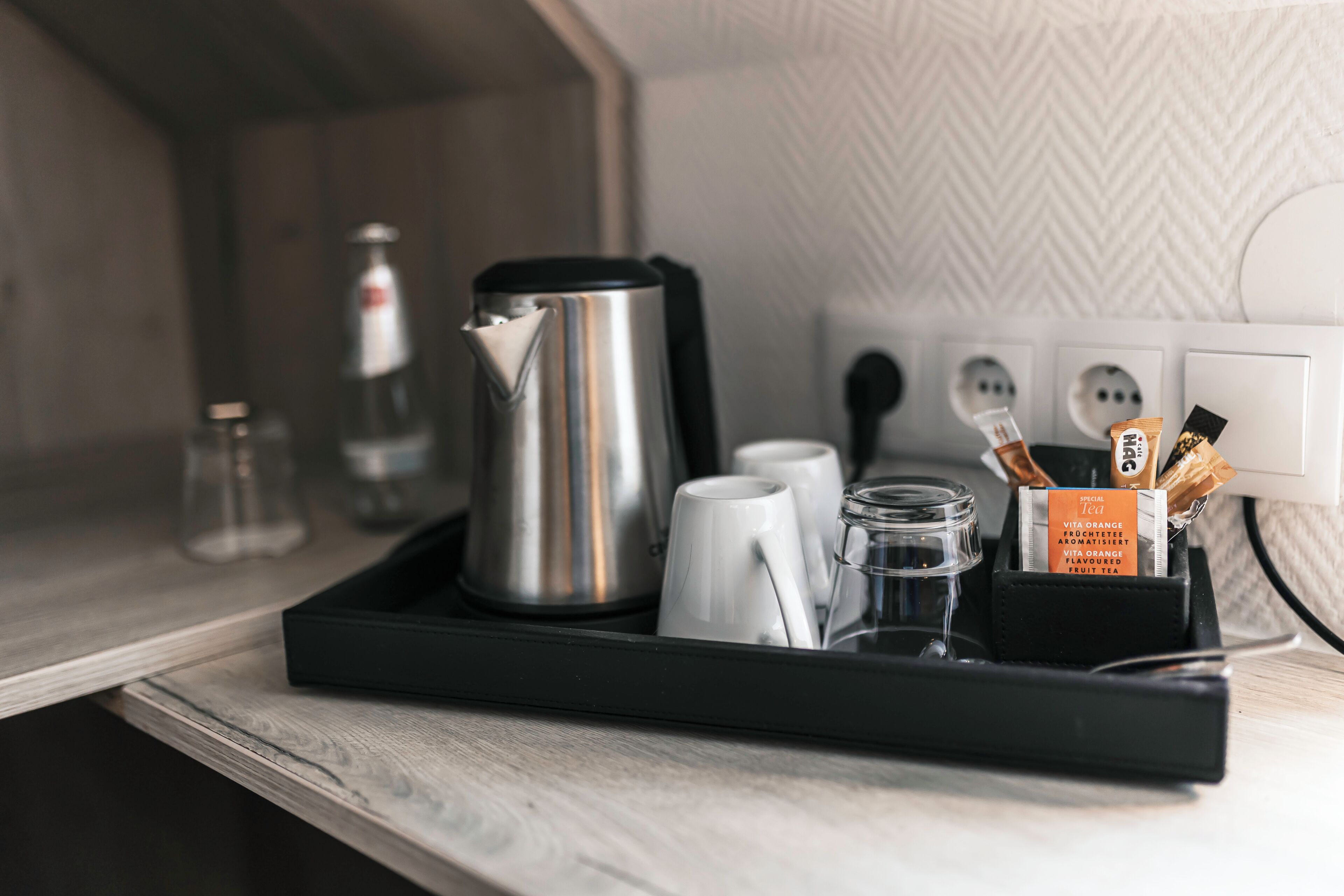 Un plateau contenant une bouilloire en acier inoxydable, des tasses blanches, un bocal en verre et des sachets de thé, le tout posé sur un comptoir en bois.