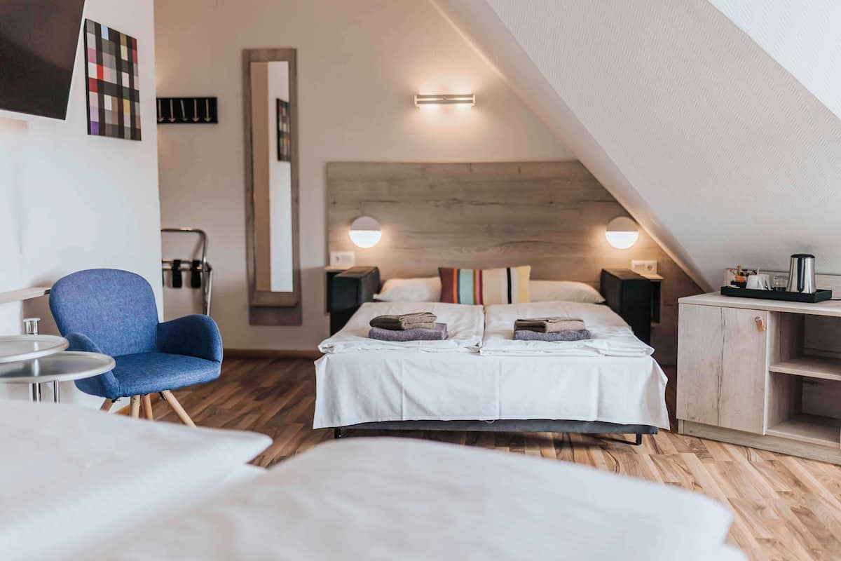Quarto moderno com teto inclinado, cama com roupa de cama branca, poltrona azul e cabeceira de madeira.