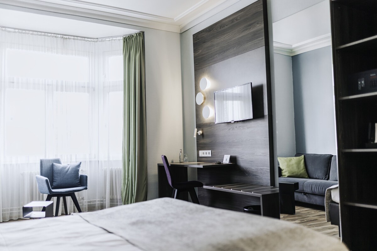 A modern hotel room with a bed, a desk with a chair, a sofa, and a TV mounted on the wall.