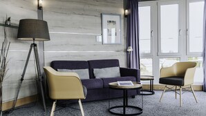 A modern hotel room with a dark blue sofa, two chairs, a small round table, a floor lamp, and a large window with curtains.