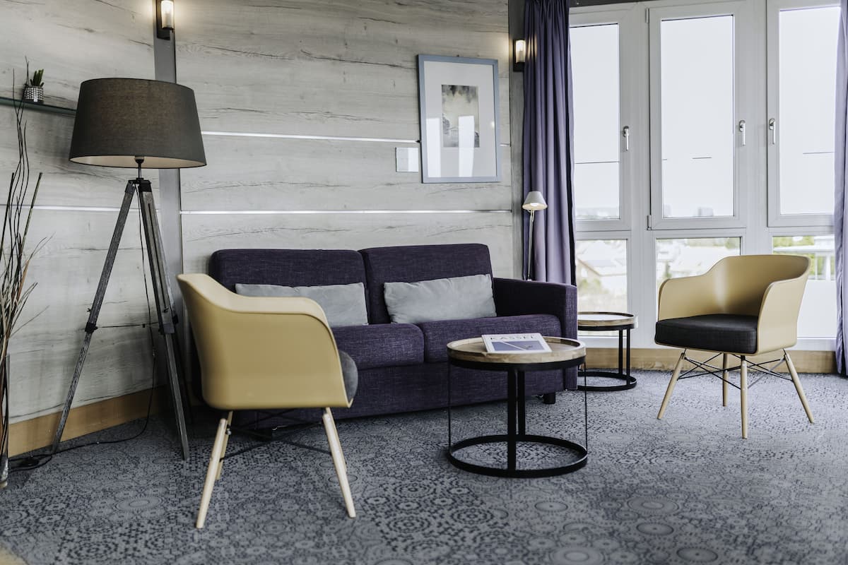 Quarto de hotel moderno com um sofá azul escuro, duas cadeiras, uma mesa redonda pequena, um abajur e uma janela grande com cortinas.
