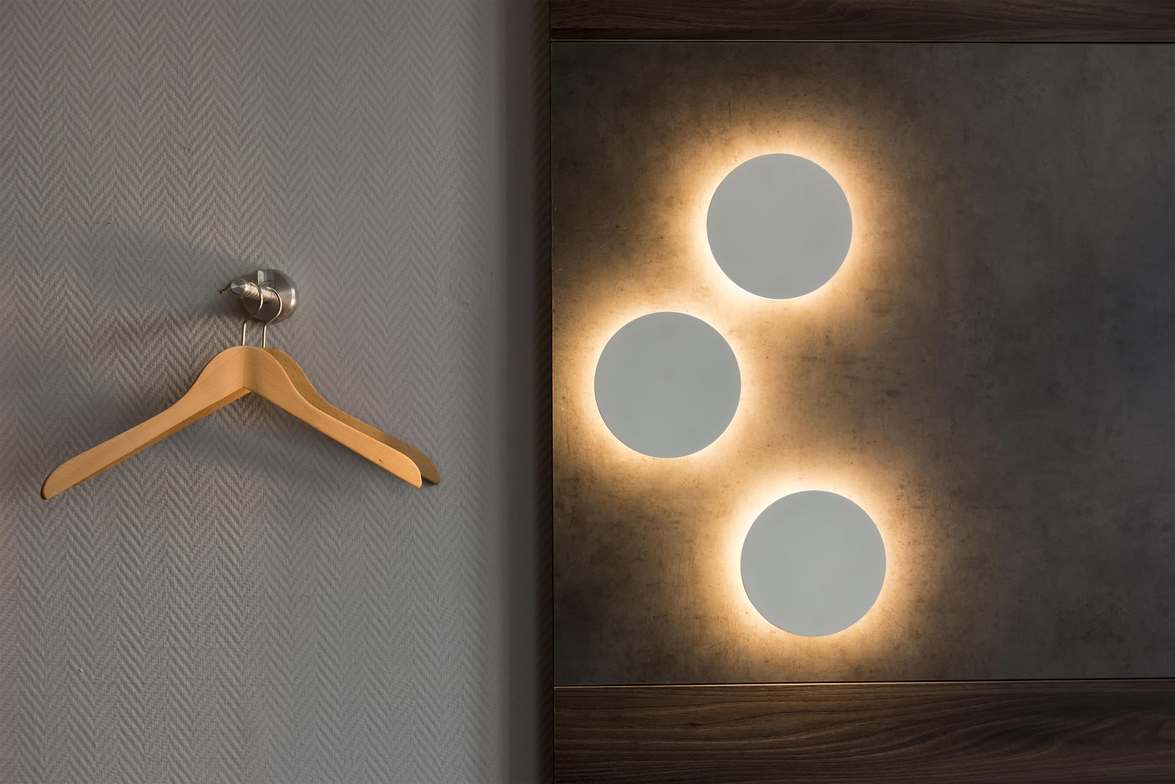 Ein hölzerner Kleiderbügel an einer strukturierten Wand, eine Wandleuchte mit drei beleuchteten Kreisen und ein Holzpaneel darunter.