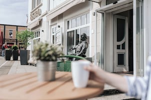 Un bistró con una estatua en el exterior, una planta en maceta sobre la mesa y una taza blanca.