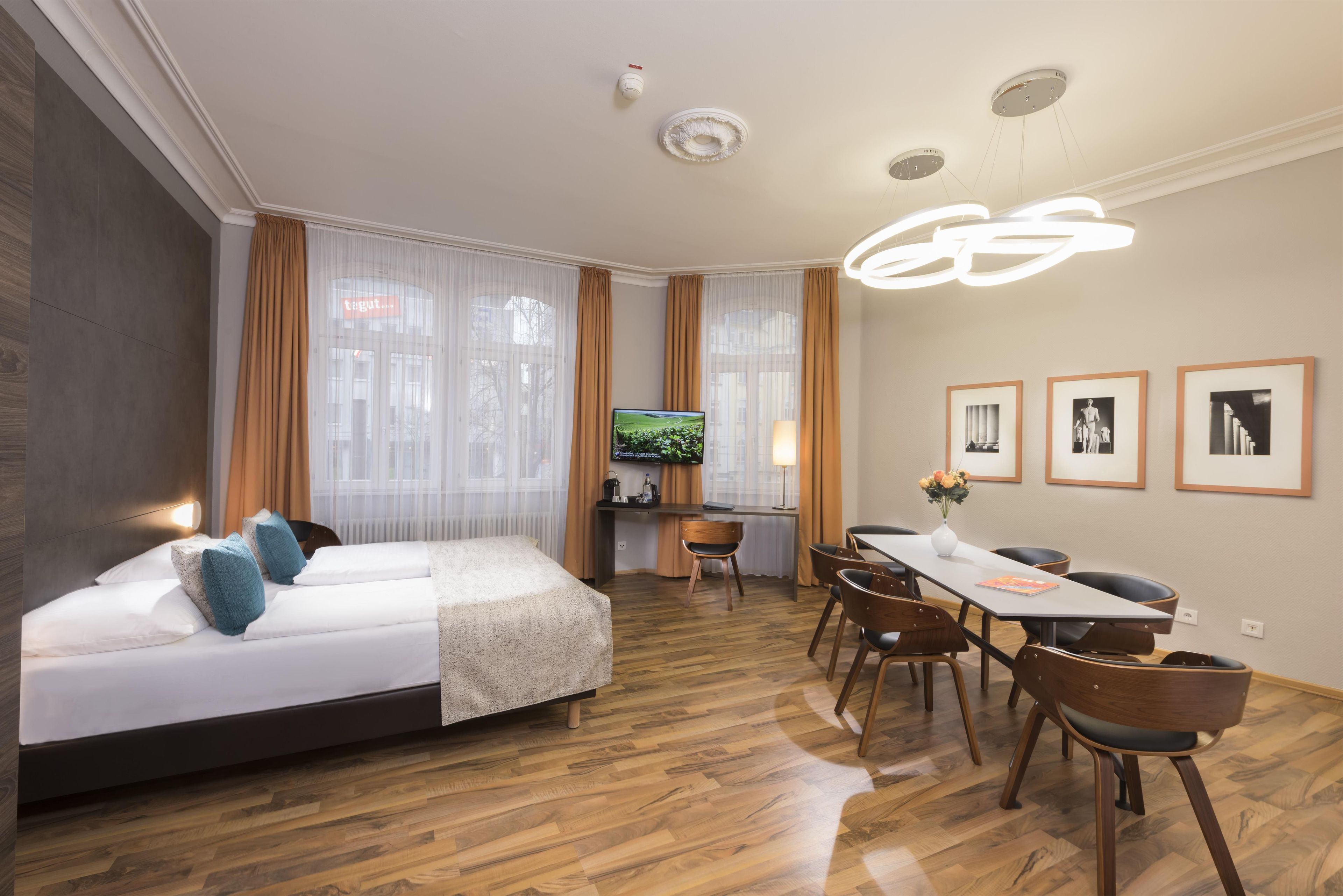 A hotel room with a bed, a desk, a dining table with chairs, a television, and framed pictures on the wall.