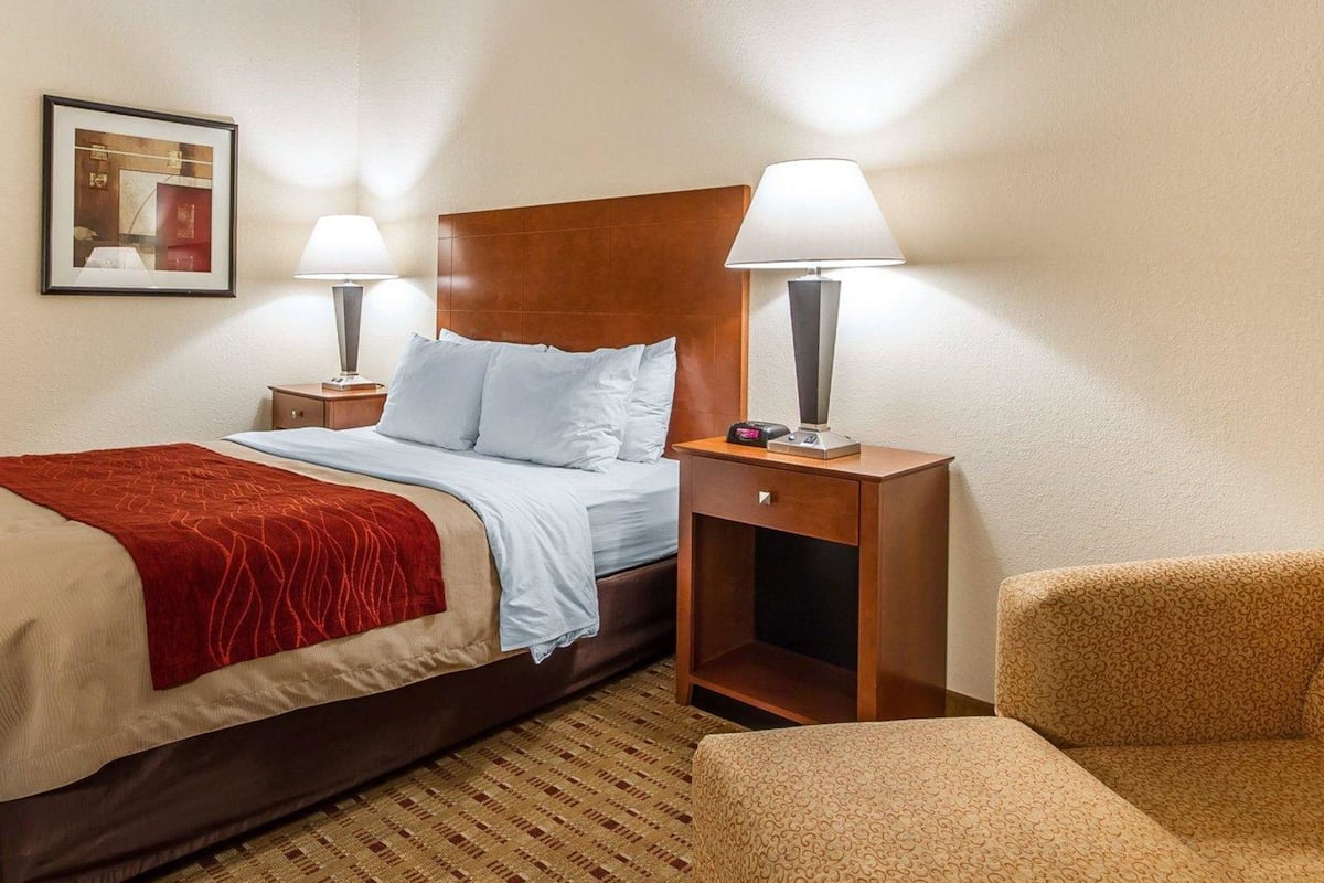A hotel room with a bed, two bedside tables with lamps, a chair, a framed picture on the wall, and a patterned rug.
