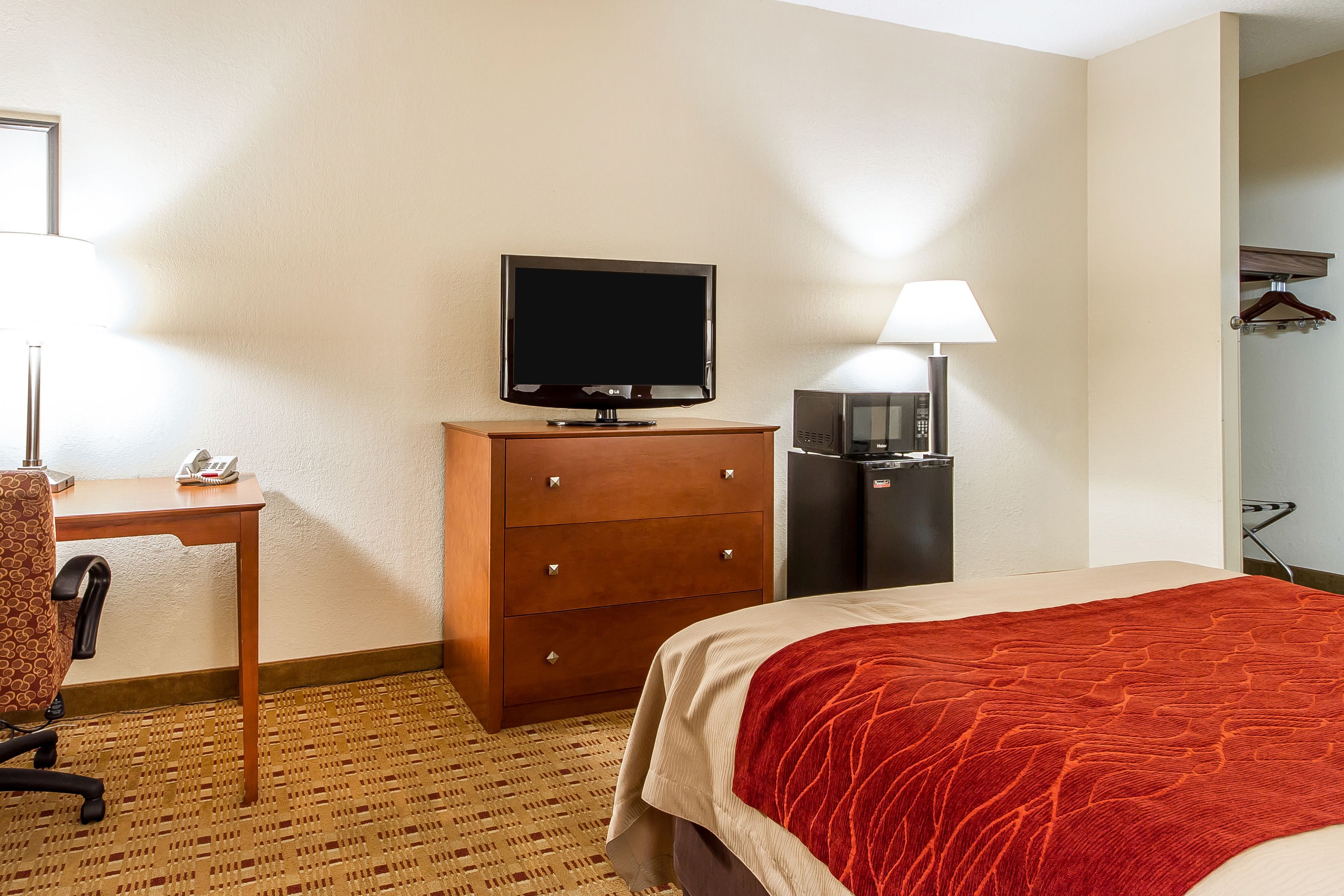 Una habitación de hotel con cama, escritorio con silla, televisión, refrigerador y lámpara.