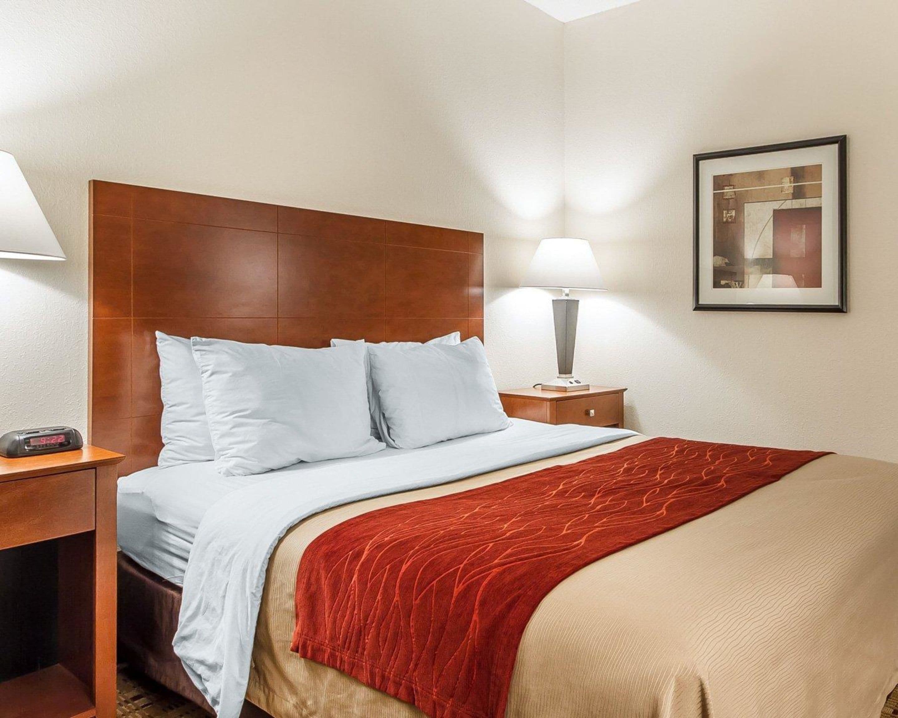 A hotel room with a wooden headboard, a bed with white and red bedding, a nightstand with a lamp, and a framed picture on the wall.