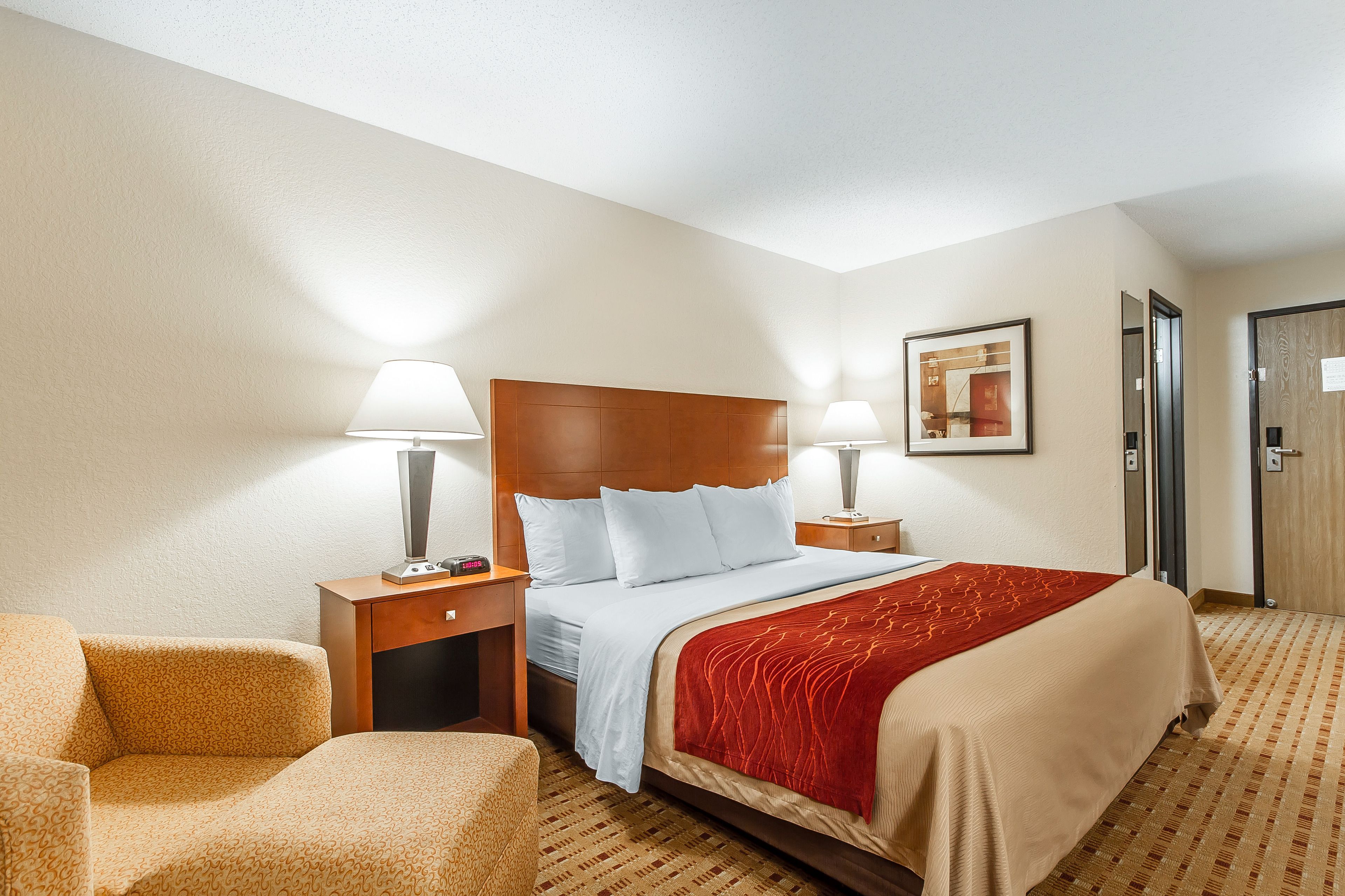 A hotel room with a large bed, two bedside tables with lamps, a chair, a framed picture on the wall, and a door.