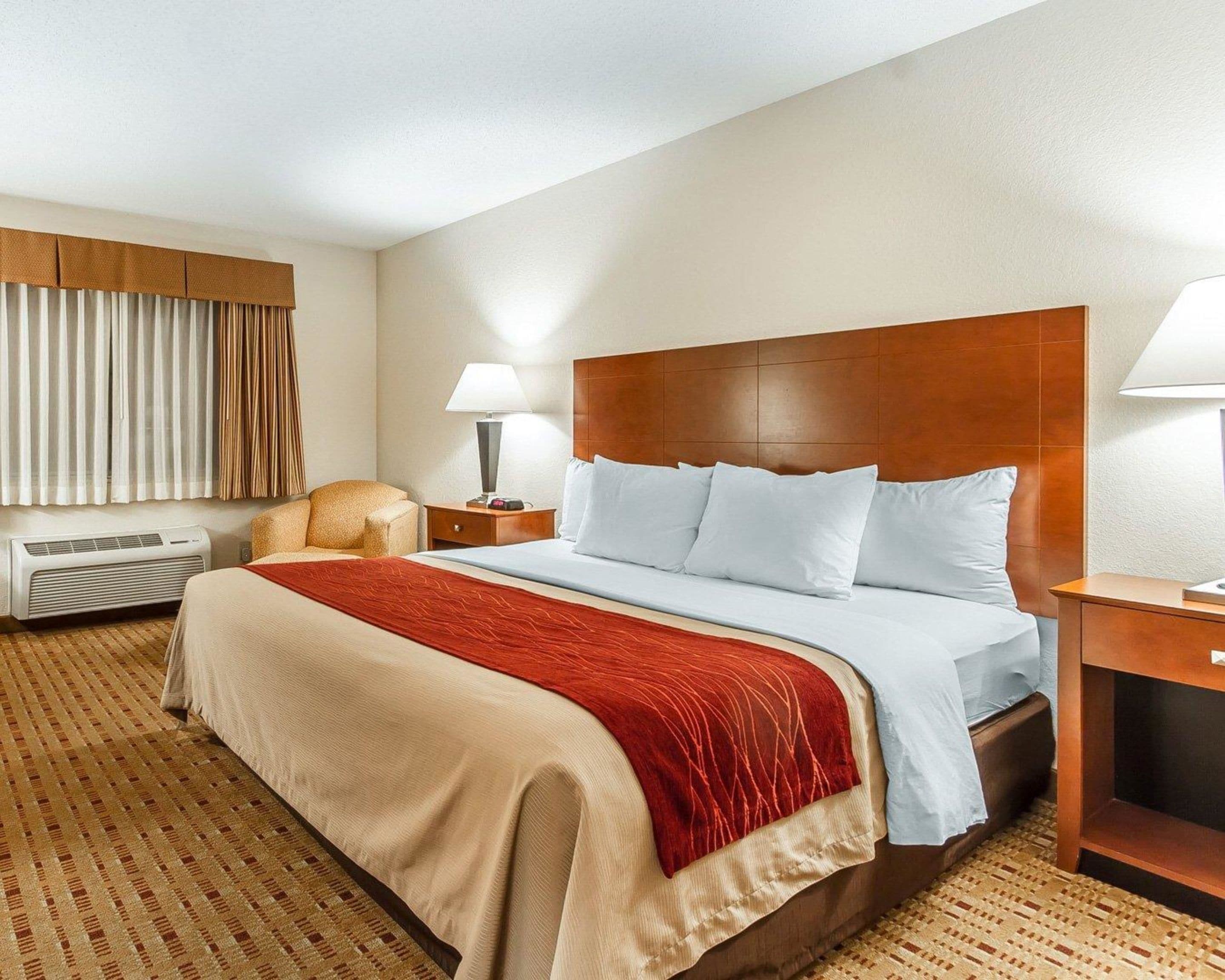 Una habitación de hotel con una cama grande, una mesita de noche con lámpara, una silla, una ventana con cortinas y aire acondicionado instalado en la pared.