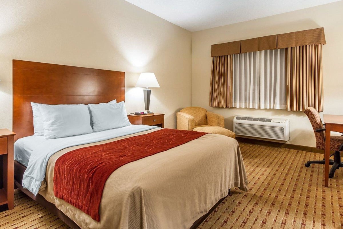 A hotel room with a bed, a desk, a chair, a lamp, and an air conditioning unit.