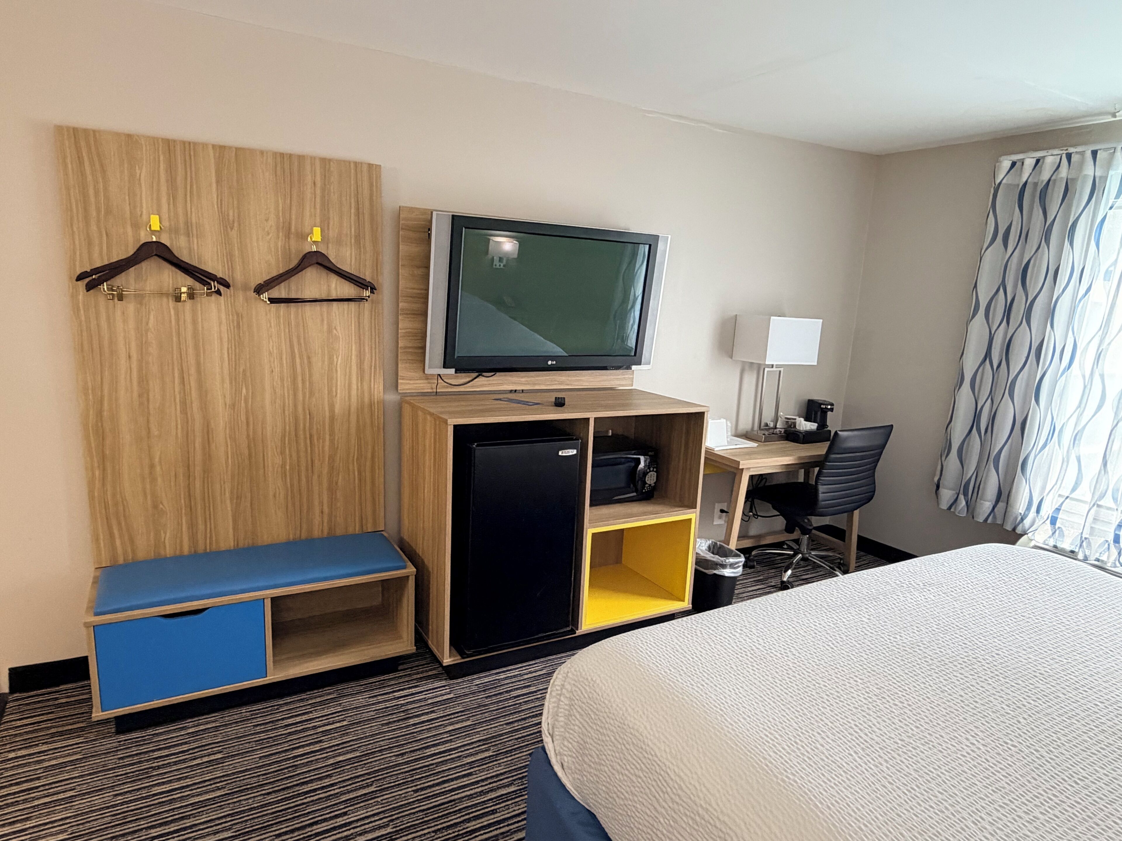 A hotel room with a bed, a desk with a computer, a television, a wardrobe, and a seating area.