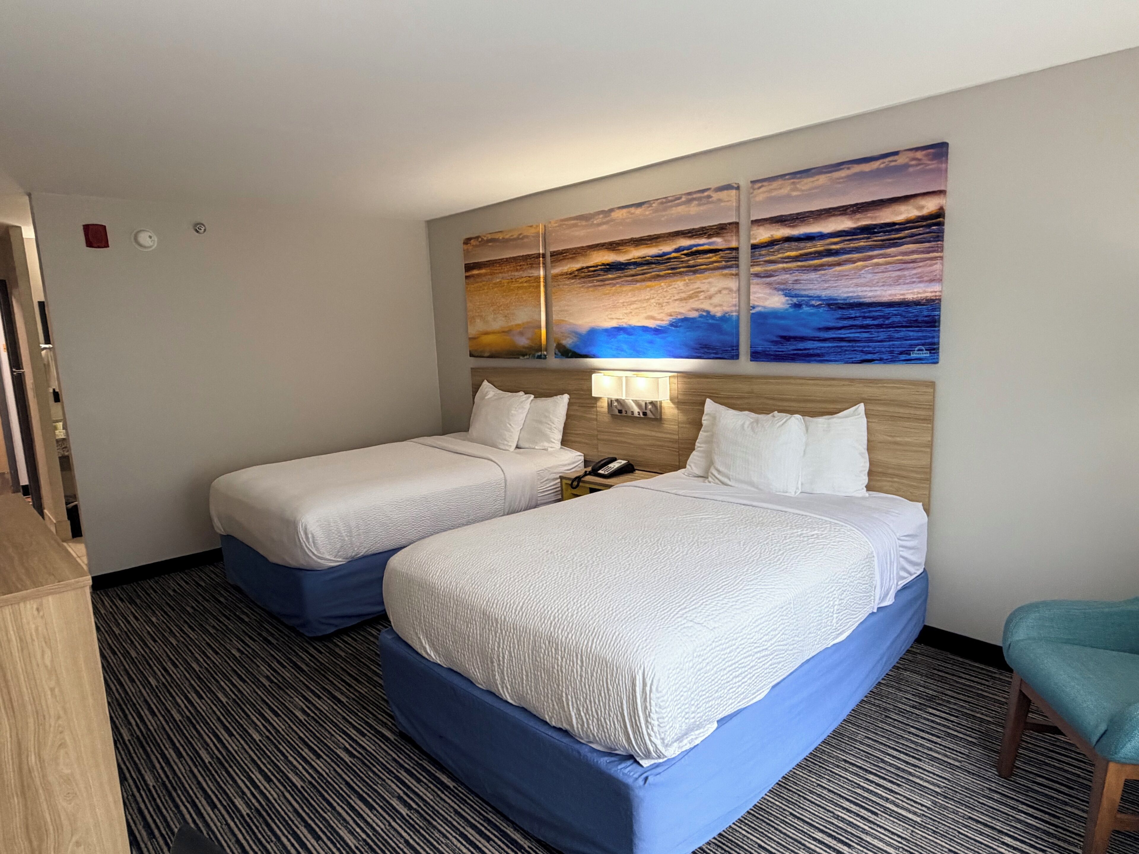 A hotel room with two beds, a nightstand with a phone, and a wall with a sunset painting.