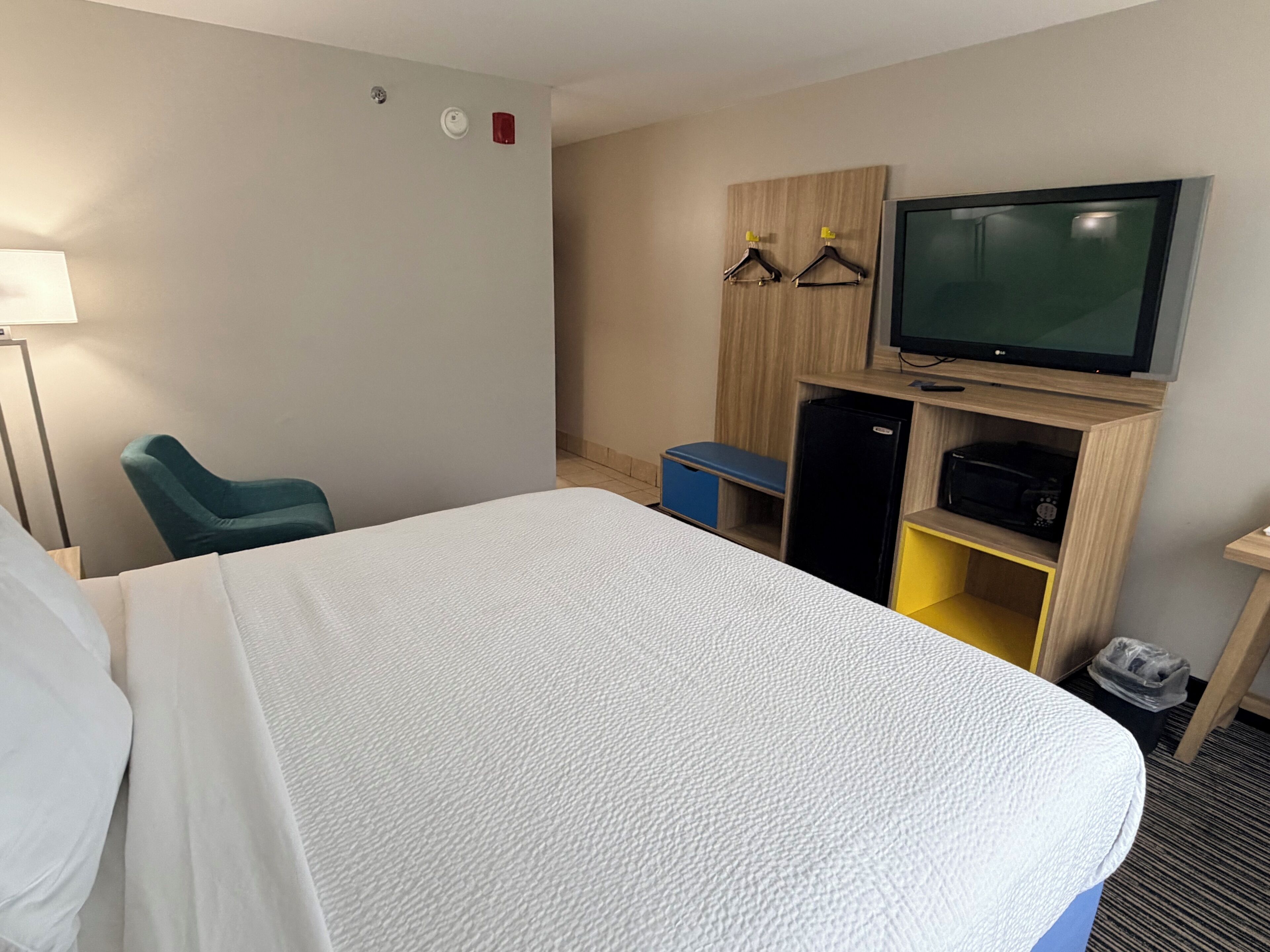 A hotel room with a bed, a television, a desk with a computer, a chair, and a small storage unit.