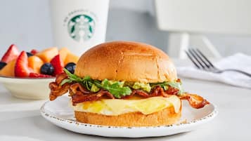 Un sandwich au bacon, à la laitue et au fromage servi dans un pain, une assiette de fruits et, au fond, une tasse Starbucks.