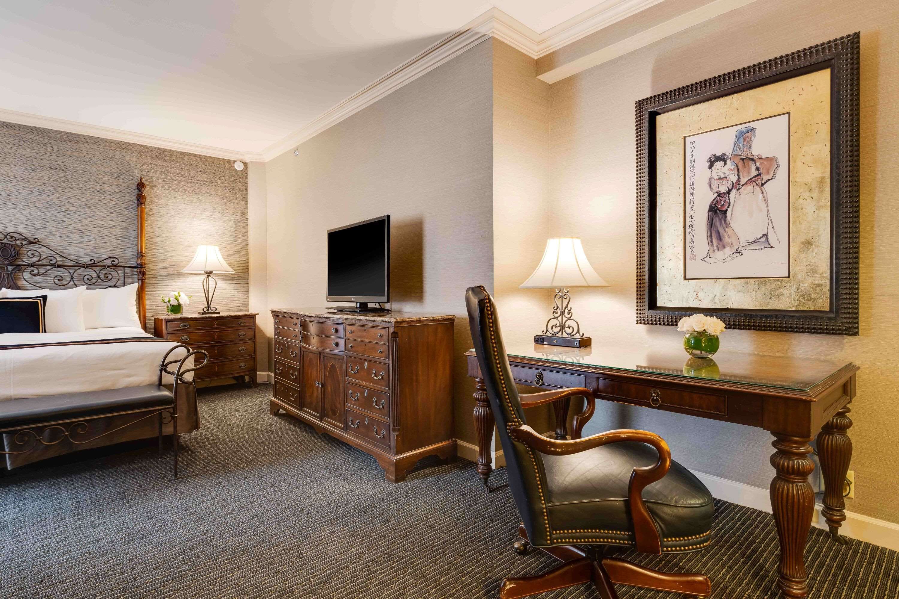 A hotel room with a bed, a desk, a chair, a dresser, a television, and a framed artwork on the wall.
