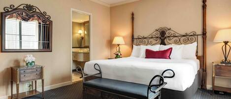 A hotel room with a large bed, a bench, a mirror, a lamp, and a small table with a vase of flowers.
