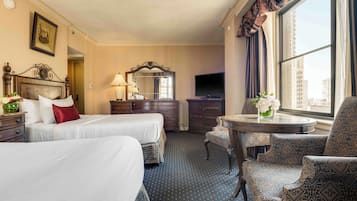 A hotel room with two beds, a desk, a chair, a TV, and a large window with curtains.