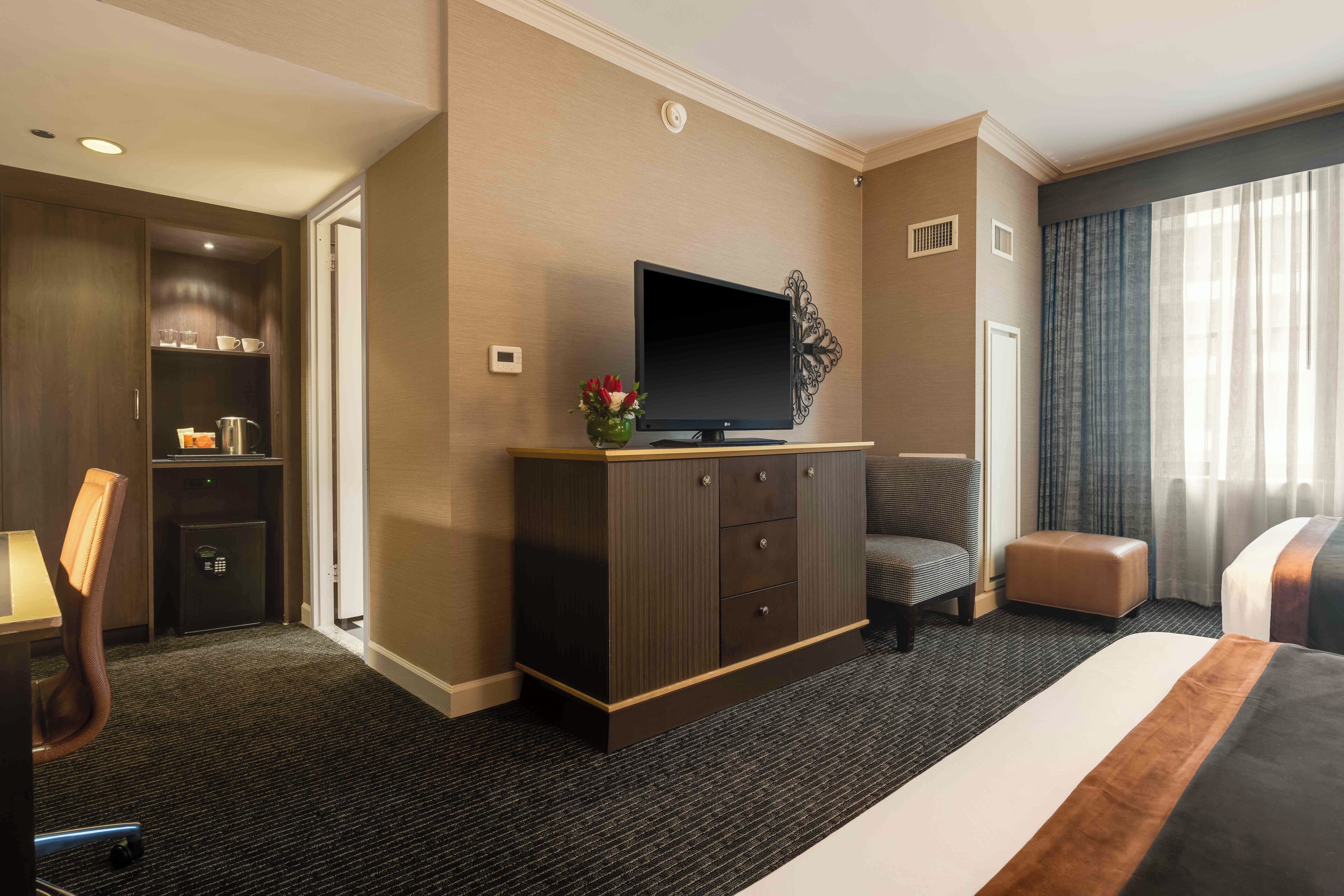 A hotel room with a large bed, a flat-screen TV on a wooden cabinet, a desk with a chair, a safe, and a small seating area.