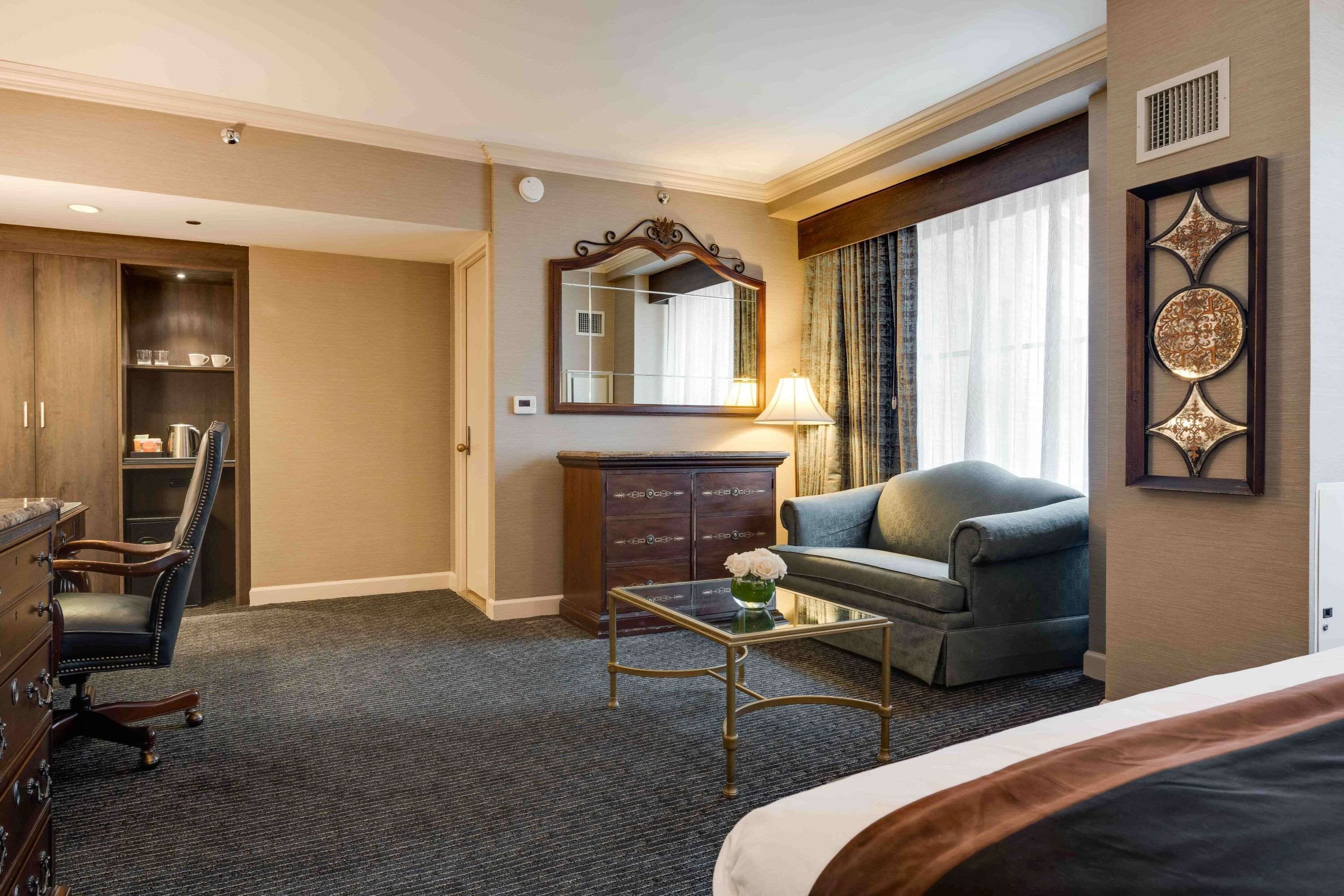 A hotel room with a bed, a desk, a chair, a sofa, a dresser, a mirror, and a window with curtains.